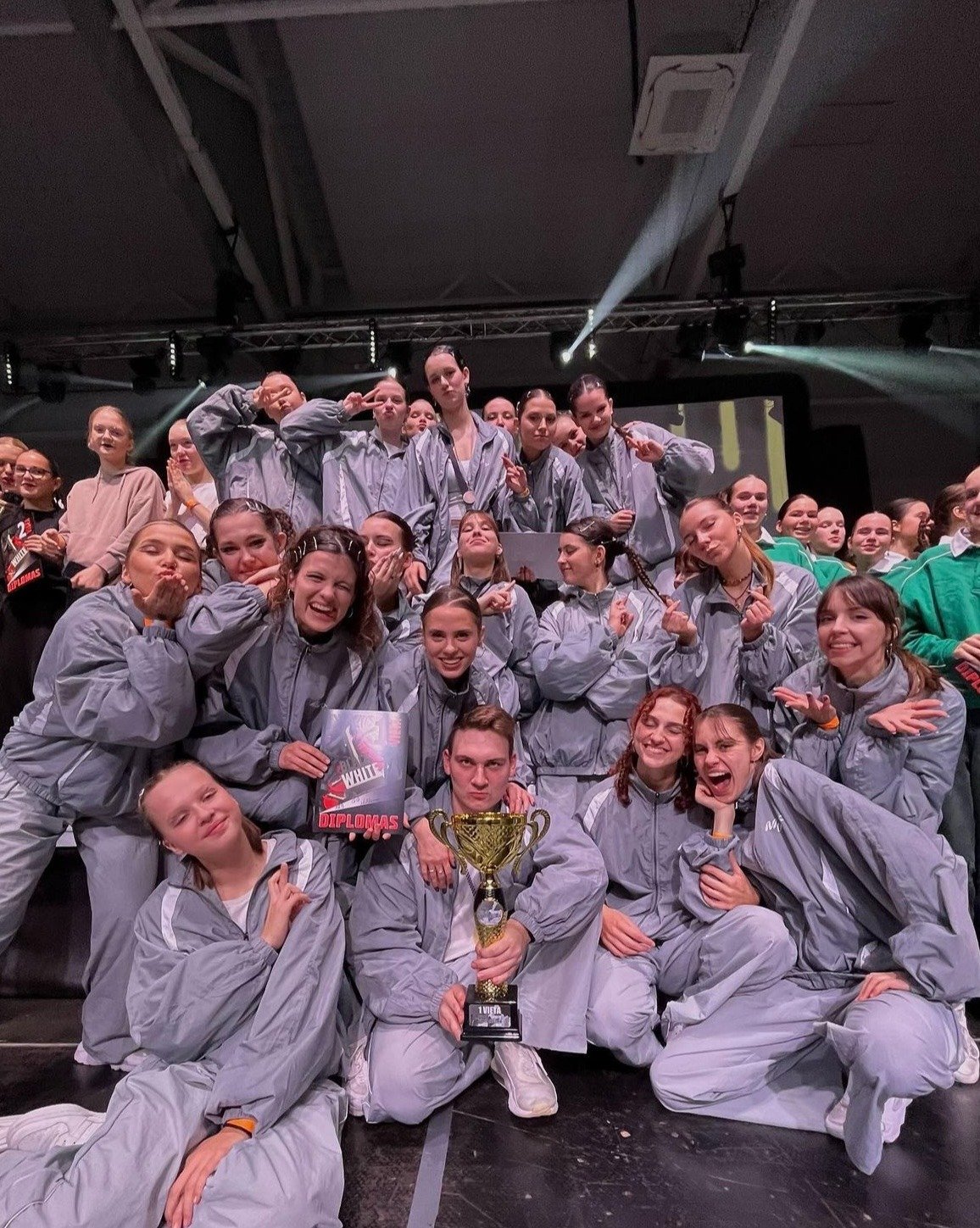 A victorious hip hop dance team in grey tracksuits celebrating with a trophy and diploma award on stage.