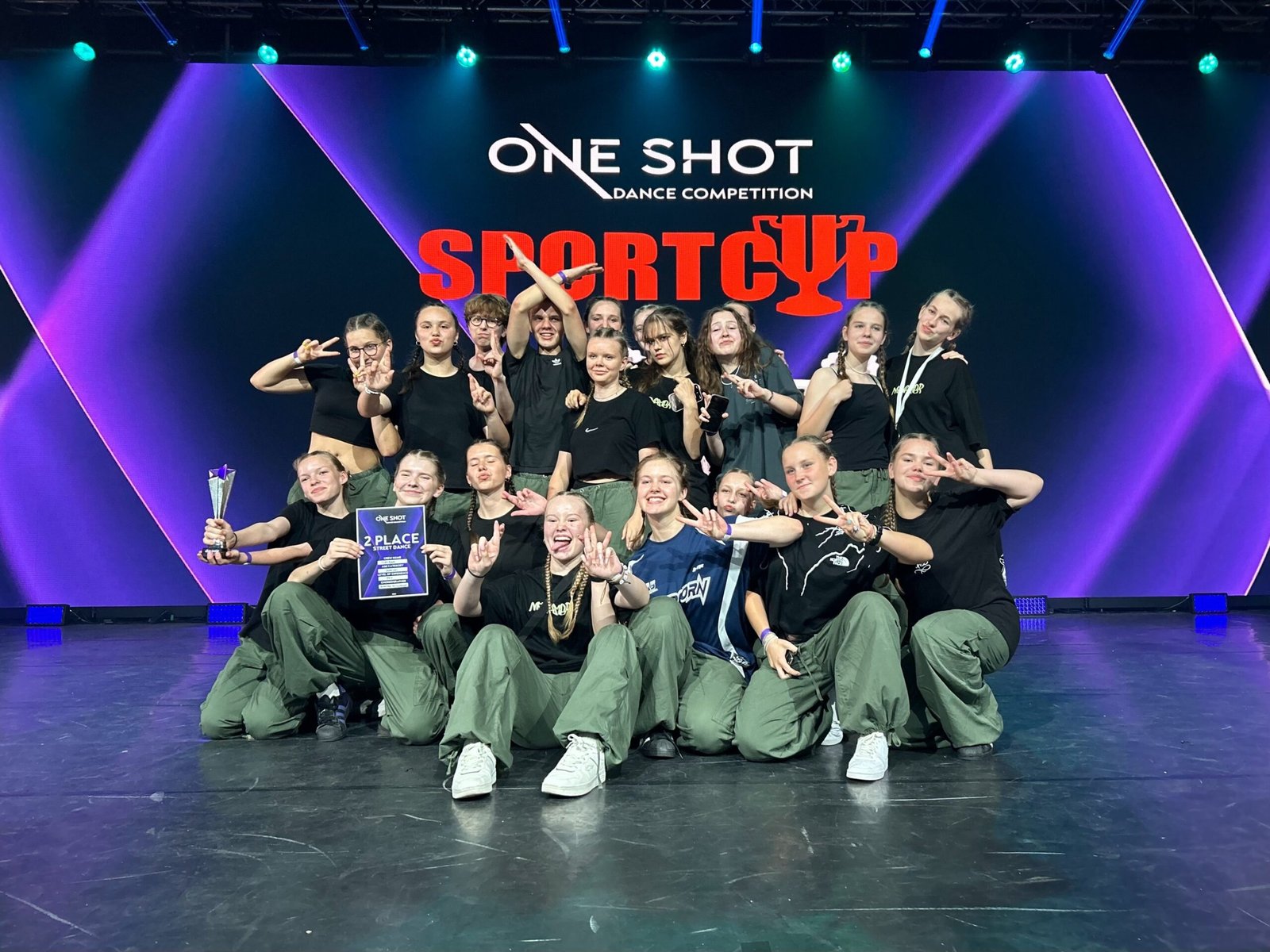 A hip-hop dance crew posing on stage with a 2nd place trophy at the One Shot Dance Competition Sport Cup.